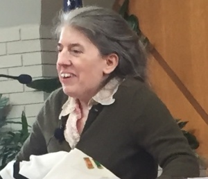 Ingrid Tischer speaking into a microphone at congregation Beth Jacob. She is a white woman with graying hair who is holding herself up with some visible effort.