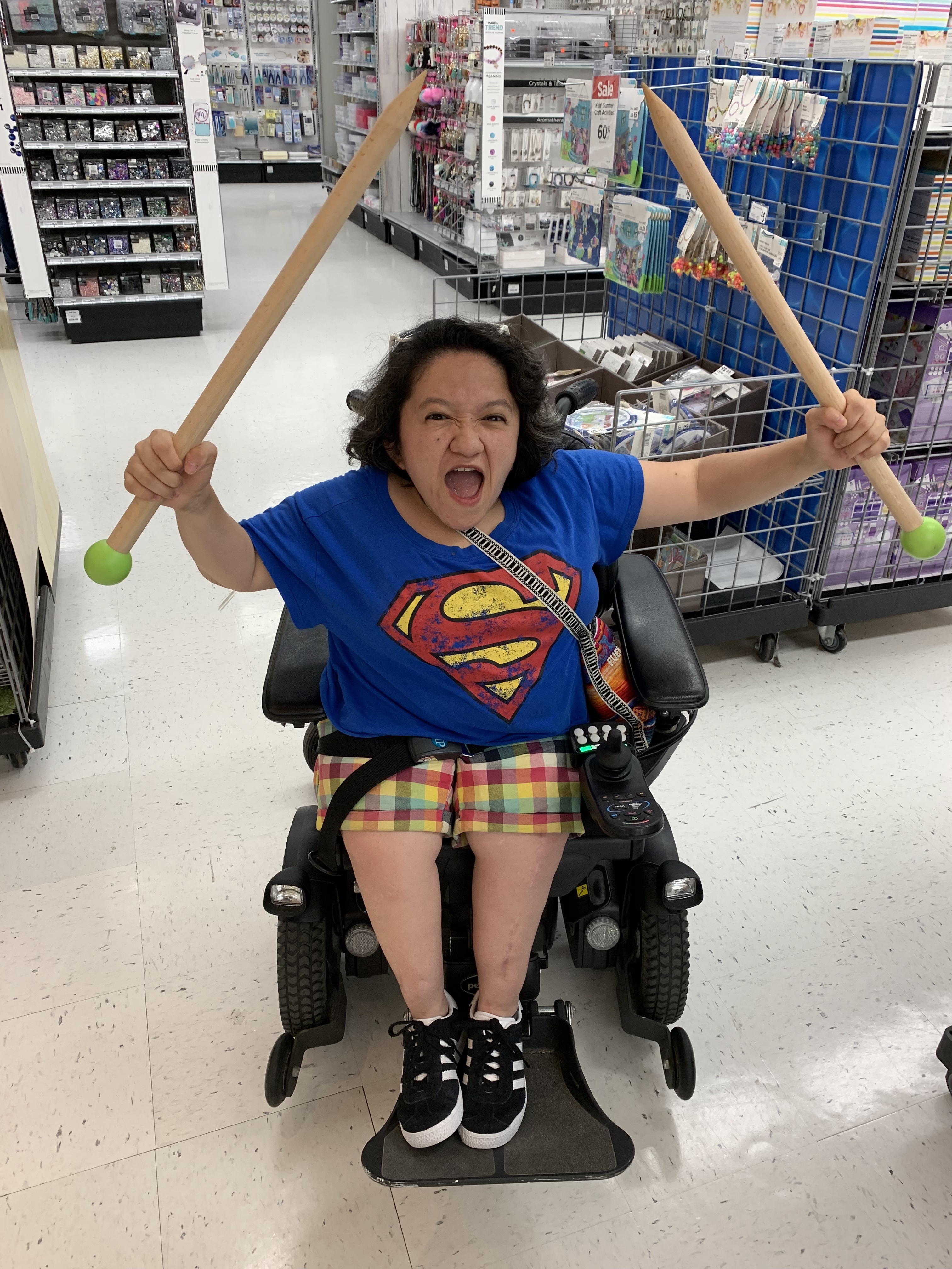Sandy Ho, an Asian American woman with dark curly hair sits in a black power wheelchair. She is wearing a blue tshirt with a superman logo, she also has brightly colored plaid shorts on and black sneakers. Her facial expression is one of outraged protest and in both upraised fists she holds oversized wooden knitting needles.