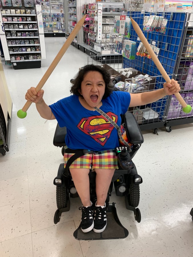 Sandy Ho, an Asian American woman with dark curly hair sits in a black power wheelchair. She is wearing a blue tshirt with a superman logo, she also has brightly colored plaid shorts on and black sneakers. Her facial expression is one of outraged protest and in both upraised fists she holds oversized wooden knitting needles.