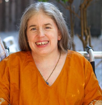 Ingrid,a middle-aged White woman with long graying hair, sitting in a wheelchair and smiling